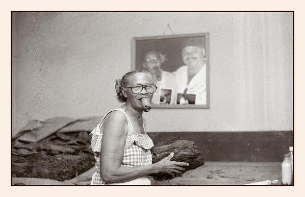 Ältere cubanische Frau mit einer dicken selbstgedrehten Zigarre im Mund und einem Bündel Tabakblätter in der Hand. Im Hintergrund liegen Tabakblätter auf einem Tisch. Ein Bild hängt im Hintergrund an der Wand. Foto aus der Partagas-Manufaktur in Havanna.