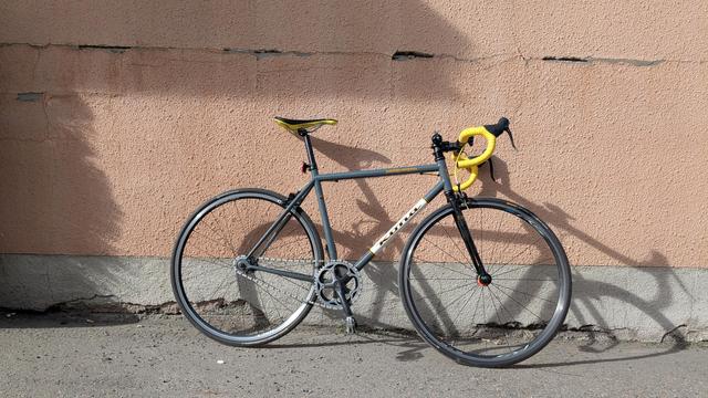 A Kona Paddywagon single-speed bicycle leaning up against a wall. The pristine yellow bar tape is glistening. A sharp bike-shaped shadow is cast on the wall by the strong sunlight.