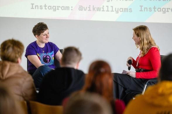 Two women sitting on chairs are talking to each other. They are in front of a screen with Instagram handles. There are people sitting behind them listening.