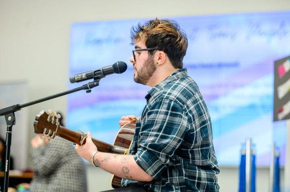 A young man with tattoos on his arms is singing into a microphone while playing an acoustic guitar. He is wearing a plaid shirt and glasses. The background is a light blue wall.