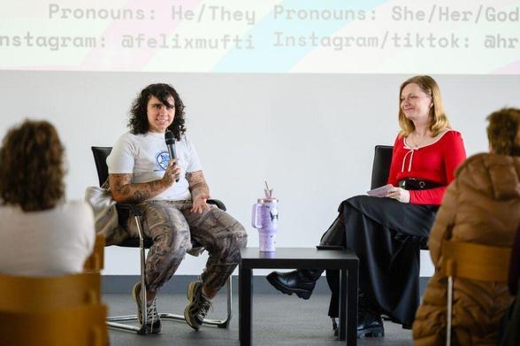 Two people sitting in chairs and talking. One person is holding a microphone. There is a screen behind them.