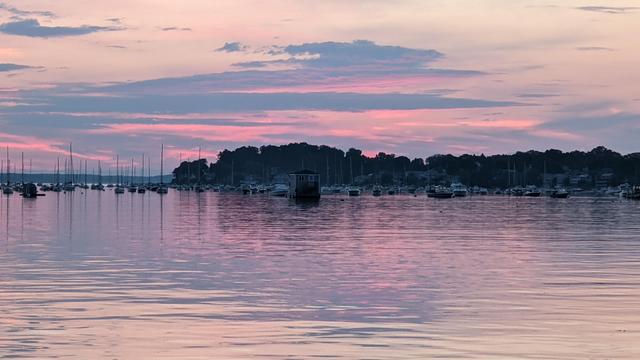 Historical Documentation in the form of a photograph of a rising sun over the ocean bay at the Historic Pioneer Village in Forest River Park, Salem, MA.  The sky is filled with vibrant colors of pink, orange, and yellow, with the sun rising in the background. The water is calm and reflects the colors of the sky. In the center of the image, there is a small boat docked at a pier with several sailboats and yachts moored in it. The harbor is surrounded by trees and buildings, and the horizon line is visible in the distance. The overall mood of the photograph is peaceful and serene.