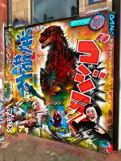 Mural of Godzilla surrounded by Japanese writing and people screaming to get away. Very bright and bold colours, with Godzilla in the center of the piece. By DANK aka Dan Kitchener, in East London. 