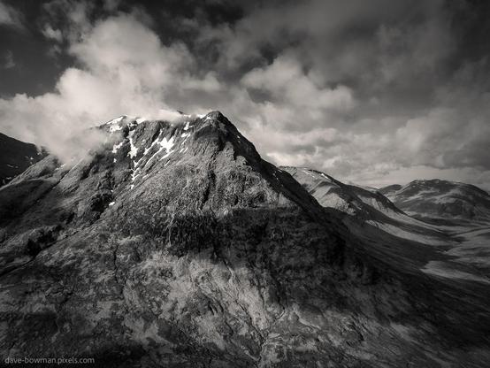 A photograph of Sron na Creise at the entrance to Glen Etive in the Scottish Highlands, captured from above. The rugged slopes rise steeply, their contours partially veiled by drifting clouds. Patches of sunlight filter through, casting shifting patterns across the mountainside and highlighting the landscape’s dramatic texture.