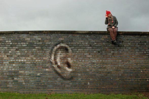 Conferences That Work meeting design: A photograph of a person sitting on top of a high brick wall with their hand cupped around their right ear. The wall displays an ear distorted out of the brick surface.
