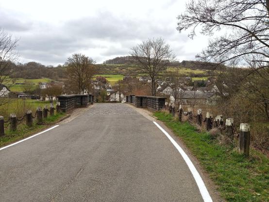 View towards a neat historical bridge with cobblestones and bollards to both sides of the road. In the background village Ettersdorf and behind a hill with meadows, scrubs and on its top a small forest. Some trees surround the road in the foreground. Cloudy sky in early spring.