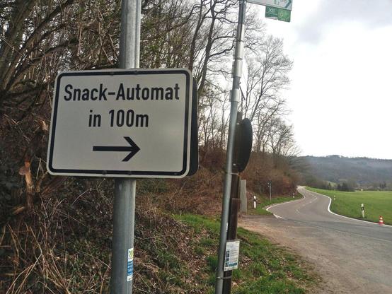 In the foreground a sign towards a snack vending machine. In the background a road winding its way through the valley. Left scrub, right meadow. In the background a hill. Weather is cloudy in early spring.