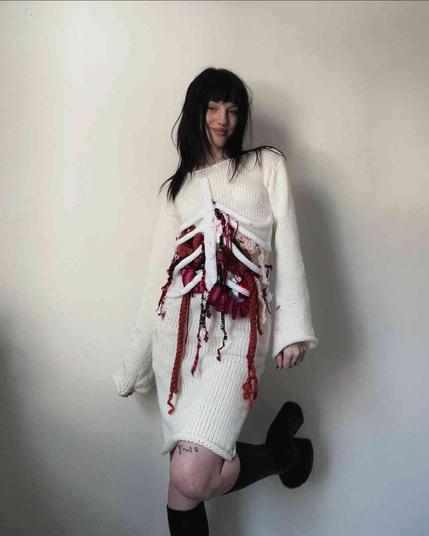 Woman wearing a white crocheted sweater dress with a crocheted design depicting a rib cage and internal organs, in red and white yarn.