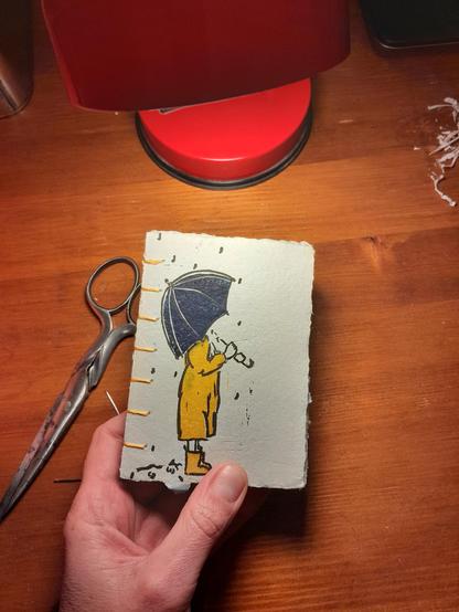 Carnet en papier artisanal, reliure copte. Linogravure représentant un.e enfant en bottes et ciré jaune sous un parapluie bleu