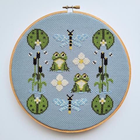 Finished cross stitch project with swampy elements such as frogs, dragonflies, lily pads and reeds
