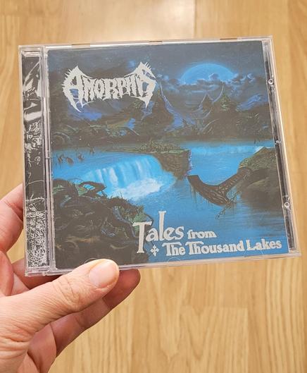 A CD album held by a hand on the left side, beige and orange flooring can be seen underneath. Album cover is mostly done in blues and depicts a fantastical landscape, dominated by a huge lake that is surrounded by gnarly trees and tree roots. There are some in the water almost at the left edge, too. A big island with more trees on it splits the lake and there are cliffs forming waterfalls on both sides of said island. Another rock is seen on the right side, and a big anchor like object with decorations carved in it is leaning on the rock (it's an ukonvasara - a long handle ending in the middle of a crescent shaped axe or hammer). It is attached to a chain that disappears in the water and is dripping with algae. The lake is surrounded by very pointy mountains, above which the night sky is full of fluffy clouds and a large moon. Band name Amorphis is left of the moon as a scraggly logo. Album name Tales from the Thousand Lakes is above the lower edge in a more print friendly font.