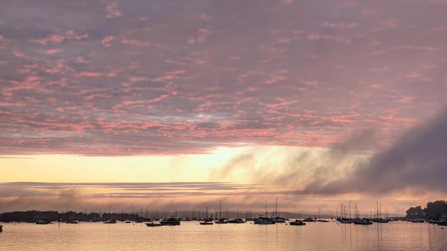 Historical Documentation in the form of a photograph of a rising sun over the ocean harbor in Salem, MA. The sky is filled with vibrant colors of pink, orange, and yellow, with the sun rises in the background. The water is calm and reflects the colors of the sky, creating a mirror-like effect. In the foreground, there are several sailboats anchored in the harbor, with their sails billowing in the wind. The harbor is surrounded by trees and hills, and the horizon line is visible in the distance. The overall mood of the image is peaceful and serene, almost magical.