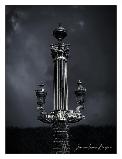 The image features an elegantly designed street lamp, striking against a dark, moody sky. The column, characterized by its fluted structure, is crowned with ornate decorations and topped by a large lamp, which adds to its grandeur. Two smaller lamps are positioned symmetrically on either side of the column's base, enhancing its regal presence.
The monochromatic color scheme emphasizes contrasts, creating a dramatic effect that imbues the scene with a sense of historical significance. The textures of the stone and metal work add depth, inviting viewers to appreciate the craftsmanship involved.
Symbolically, the column could represent enlightenment or guidance, traditionally associated with lampposts, illuminating the path ahead. The dark clouds in the background may evoke a sense of foreboding or introspection, juxtaposing the illumination provided by the lamps against the encroaching gloom.
Overall, the image captures a moment of stillness, inviting contemplation on themes of history and resilience amidst adversity.