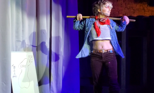 A photo of Austin Tacious dressed as a cowboy with velvet riding pants, a jean shirt, a mesh shirt under that, and a bandanna. He is standing by a board with a diagram that reads "Me (parked)," "Lake," and "SF (80 M)," holding a meter stick, and looking forlorn.