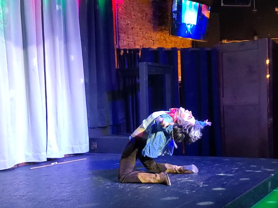 Another photo of Austin Tacious, dressed as a cowboy, standing on his knees and bending over backwards to face the audience with arms out to either side.