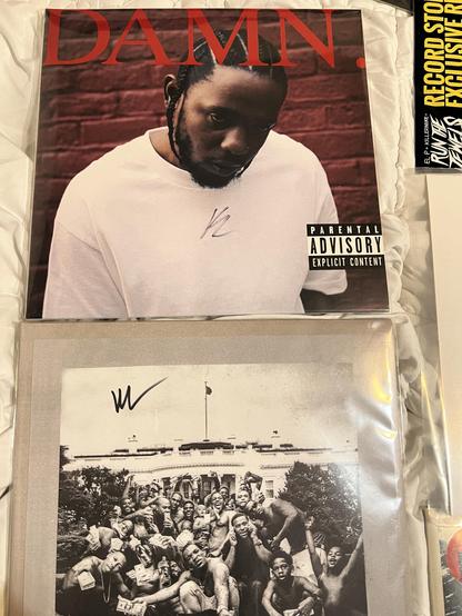 The image features two vinyl records by Kendrick Lamar. The top record displays the album "DAMN." with a close-up of Lamar on the cover, and it is signed. The bottom record features a black-and-white photo of Lamar with a group of people, the TPAB cover. 