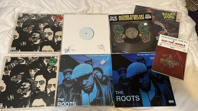 A collection of vinyl records arranged on a white sheet. Featured are albums by The Roots, Killer Mike, and Run The Jewels, including signed editions and a Record Store Day exclusive release.