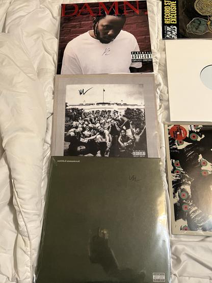 A collection of vinyl albums displayed on a white bedspread. The top album is "DAMN." by Kendrick Lamar, featuring a photo of the artist. Below it is "To Pimp a Butterfly," showcasing a group of people in front of the white house. The last is untitled unmastered, a forest green plain cover. 