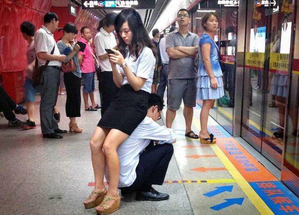 Platform in a metro station. A man is sitting on the ground, a woman sitting on his back watching at her phone.