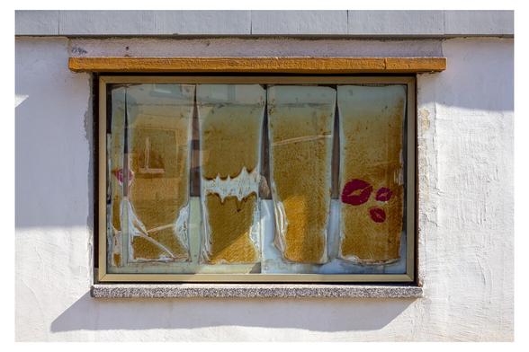 Motiv:
Ein Fenster mit vergilbten, beschädigten Sichtschutzfolien. Drei rote Lippenabdrücke befinden sich auf der rechten Scheibe. Das Fenster ist in eine verputzte Fassade eingelassen.

Stimmung & Wirkung:
Das Bild wirkt dokumentarisch und leicht ironisch. Der Kontrast zwischen Verfall und der grafischen Note der Lippen erzeugt Spannung. Es vermittelt Spuren von Zeit, Nutzung und menschlicher Intervention.

Technische Details:
Farbe: Vorherrschende Töne sind Beige, Ocker, Weiß und Rot. Die Farben wirken natürlich mit deutlicher Sättigung.
Schärfentiefe: Tiefenschärfe vollständig auf das Fenster fokussiert, Hintergrund entfällt aufgrund flacher Perspektive.
Lichtverhältnisse: Harte Lichtsituation mit direkter Sonneneinstrahlung von links, deutliche Schattenbildung an der Fassade.