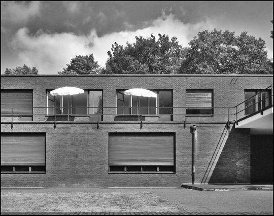 Ein modernes Backsteingebäude mit großen Fenstern und zwei weißen Sonnenschirmen auf einem Balkon. Die Szene ist in Schwarz-Weiß dargestellt, mit einem einfachen architektonischen Design und umgeben von Grün und Wolken am Himmel.
-
A modern brick building with large windows and two white umbrellas on a balcony. The scene is depicted in black and white, featuring a simple architectural design and surrounded by greenery and clouds in the sky.