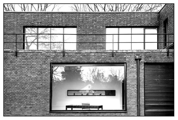 Ein minimalistisches architektonisches Äußeres mit einem Backsteingebäude mit großen Fenstern. Ein markantes Fenster zeigt einen Tisch und Dekorationsgegenstände im Inneren, die Bäume außen reflektieren. Das Bild wird in Schwarzweiß präsentiert.
-
A minimalist architectural exterior featuring a brick building with large windows. One prominent window displays a table and decorative items inside, reflecting trees outside. The image is presented in black and white.