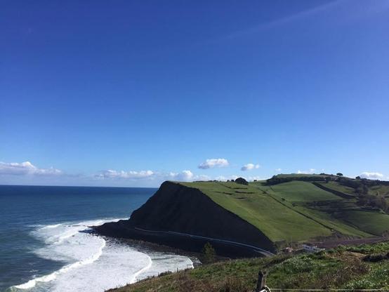 
Un acantilado oscuro y rocoso se encuentra junto a un océano azul profundo, con olas blancas rompiendo en la orilla. La cima del acantilado está cubierta de exuberante vegetación verde, que desciende en suaves colinas hacia el mar. El cielo es un azul claro y despejado, con algunas nubes dispersas. La escena evoca una sensación de tranquilidad y belleza natural.