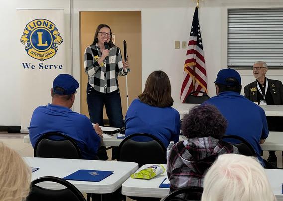 a photo of aftersight employee standing and speaking in front of a group of people. The Lions club logo can be seen in the upper left. 