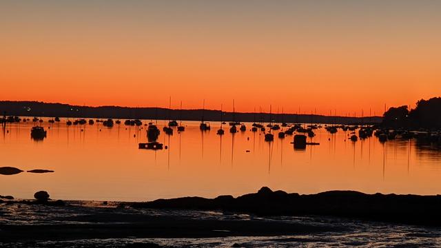 Historical Documentation in the form of a photograph of a rising sun over the ocean bay at the Historic Pioneer Village in Forest River Park, Salem, MA. The sky is a warm orange color, with the sun partially visible on the right side of the image. The water is calm and reflects the colors of the sky, creating a mirror-like effect. In the foreground, there are several sailboats anchored in the water, dreaming of their sails billowing in the wind. The boats are of different sizes and colors, including red, orange, and yellow. The horizon line is visible in the background, with trees and hills on the left side. The image is taken from a low angle, looking towards the horizon.