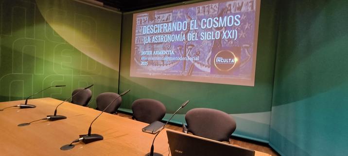Una mesa de presidencia con cuatro butacas en el Aula Magna de la UNED en Barbastro. Detrás, la pantalla con la diapo de apertura donde dice:  
DESCIFRANDO EL COSMOS
(LA ASTRONOMÍA DEL SIGLO XXI)
Y luego mi nombre y la cuenta de #mastodon por si alguien quiere
Y el año: 2025