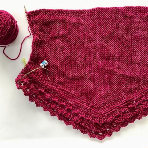 Flat lay view of a ball of dark pink yarn, and a knitting project featuring garter stitch and lace on a circular needle. It's shown on a white background.