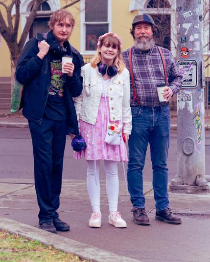 Three people stand on a street corner with a yellow building with large variously shaped windows and a poster-covered street pole beside them.  From left to right a tall man wearing dark clothes and holding a coffee cup,  a short woman with blonde hair and white tights, white shotes with pink shoelaces, a dress with a pink lower part and white upper part, white jacket, pink hair ornament and white purse, a man slightly less tall than the first with a billed cap, beard, flannel shirt, blue jeans and coffee cup.