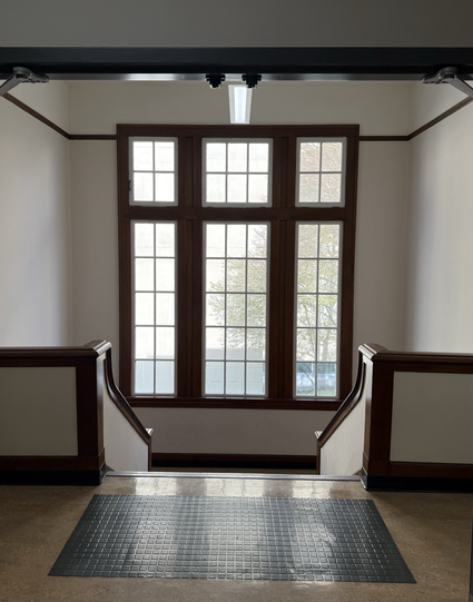 Dramatic photo of old windows from a stairwell of n old buildings. The trim is finished wood, the walls are off-white, trees are faintly visible through the windows