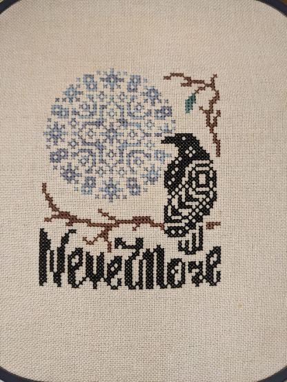 A finished cross stich of a raven sitting on a brance with a blue moon in the to left corner. Nevermore is written below the branch.