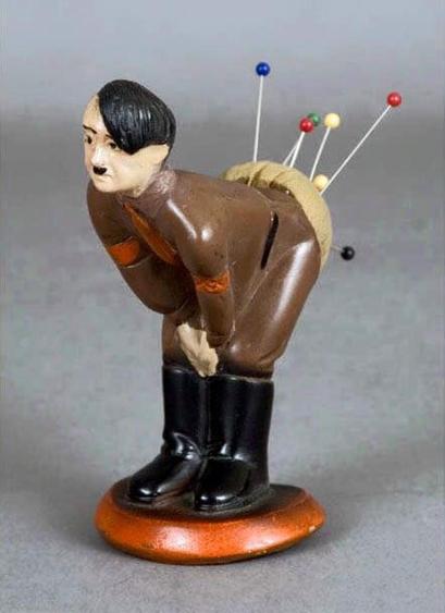 Pin Cushion Hitler, in a brown uniform and black boots, is standing, leaning forward and bending over with his hands on his knees. If you guessed that the pins are sticking out of his bum, you would be correct. 