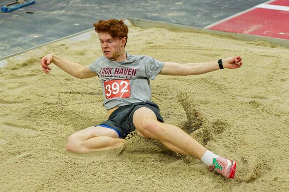 A man wearing a grey shirt with the words LOCK HAVEN UNIVERSITY and a red bib with withe letters that says 392 kicks up dirt as he lands the wrong way from the long jump