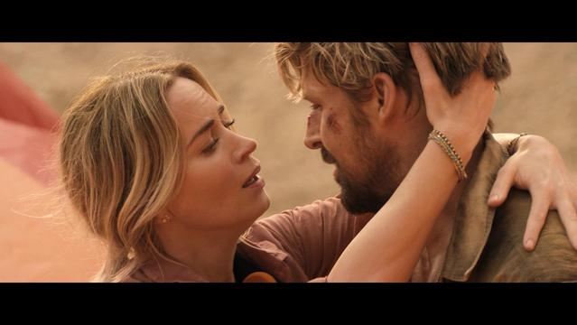 A close-up scene of two characters sharing an intense moment, showcasing emotional expressions. The female character has blonde hair and is wearing a light-colored shirt, while the male character has a rugged appearance with facial scrapes and messy hair.