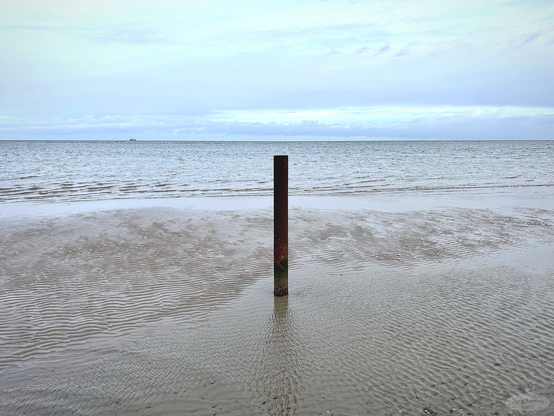 Das Bild zeigt einen rostigen Pfahl, der teilweise im seichten Wasser eines Sandstrandes steht. Im Hintergrund ist ein ruhiges Meer und ein leicht bewölkter Himmel zu sehen. Die Wellen erzeugen feine Muster im nassen Sand. Das Bild wirkt ruhig und minimalistisch.