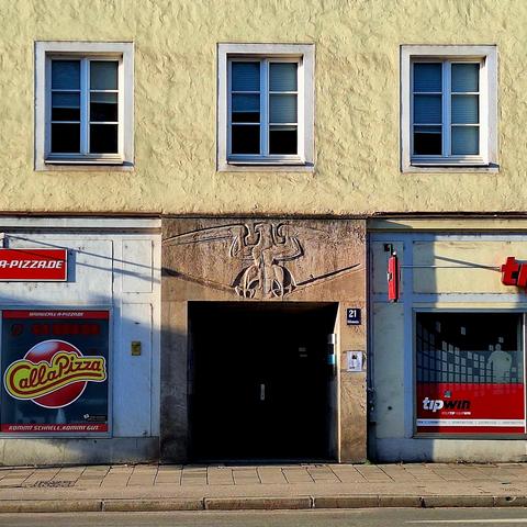 Detail of a facade of a building. Above are three simple windows surrounded by a simple plastered wall. Below are two shop windows and an entrance. Above the entrance, a large relief of a stylized eagle is visible.