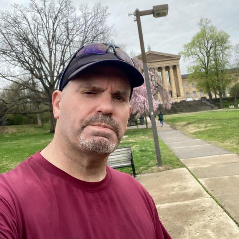 A man in a maroon shirt and black cap takes a selfie in a park with trees and a building in the background. The sky is cloudy, and there is a path and a bench nearby.