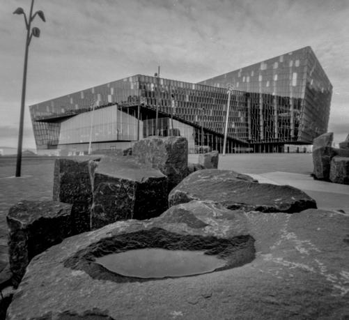 The concert hall Harpa in Reykjavik. Black and white picture taken with a pinhole film camera. In the foreground is a small water puddle on a rock.