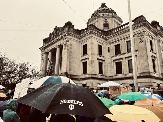 in the rain at a courthouse lawn rally, an umbrella reads "Hoosiers"