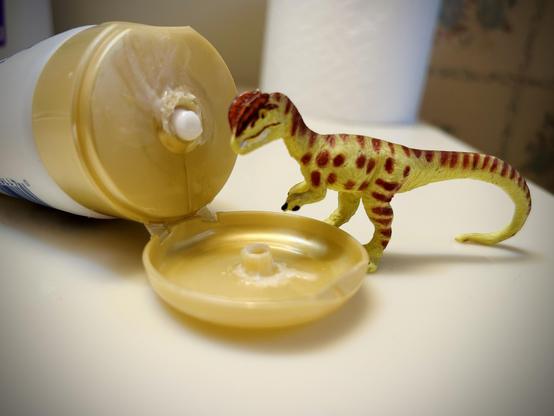 A green Dilophosaurus with brown spots stands with a bottle of Gold Bond Healing lotion in a white tube with a gold coloured cap. Both are on a white porcelain sink. A spare roll of toilet paper is in the background, taunting the viewer.