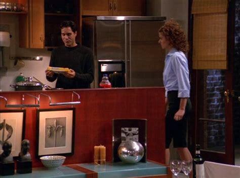 A scene from the original run of the sitcom Will & Grace showing Will & Grace in a modern apartment kitchen/living room. On the left, Will with dark hair wearing a black shirt is looking at a piece of paper. On the right stands Grace with curly hair wearing a light blue top and dark pants. They're positioned on opposite sides of a reddish kitchen island/counter. The apartment features contemporary decor with wooden cabinets, stainless steel appliances, and decorative items including a silver spherical art piece on a shelf beneath the counter.