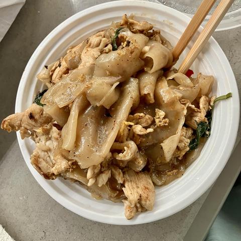 A bowl of stir-fried rice noodles with chicken, topped with green herbs. Chopsticks are resting on the side of the bowl.