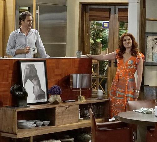 A scene from the Will & Grace set in a contemporary kitchen/dining area. On the left, Will with short hair wearing a light blue button-up shirt stands behind a wooden kitchen island holding a mug with the letter 'W' on it. On the right, Grace with long auburn/red hair is wearing a vibrant orange patterned dress with floral designs and is smiling while standing by the island. The kitchen features wood cabinetry, and the island has decorative items displayed underneath including a framed black and white photograph, a vase with purple flowers, and various other home accessories. Glass doors showing greenery outside are visible in the background.

Maybe the replacement of the silver sphere art piece and the black and white photo of Atlas holding up the world with a Google Home has symbolic meaning? Or maybe I’m just being too deep again?