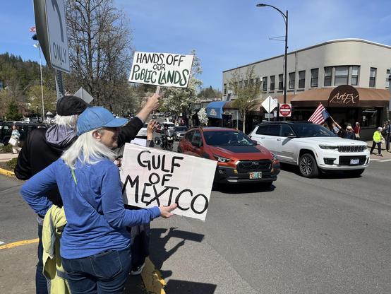 People holding signs: one says hands off our public lands, the other says Gulf of Mexico.