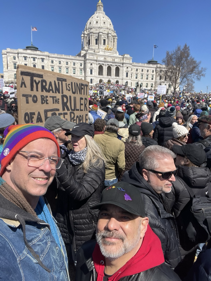 Two men take a selfie at a protest in front of the Minnesota State Capitol. One wears a rainbow-striped beanie and glasses, the other a black cap with colorful shapes. A large crowd surrounds them, holding signs like “A TYRANT IS UNFIT TO BE THE RULER OF A FREE PEOPLE.”