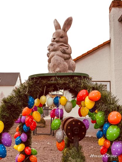 Nahaufnahme des Osterbrunnens mit Fokus auf die Hasenfigur: Ein großer Hase hält ein Hasenkind liebevoll im Arm. Im Hintergrund sind bunte Ostereier und die Girlanden zu sehen.
