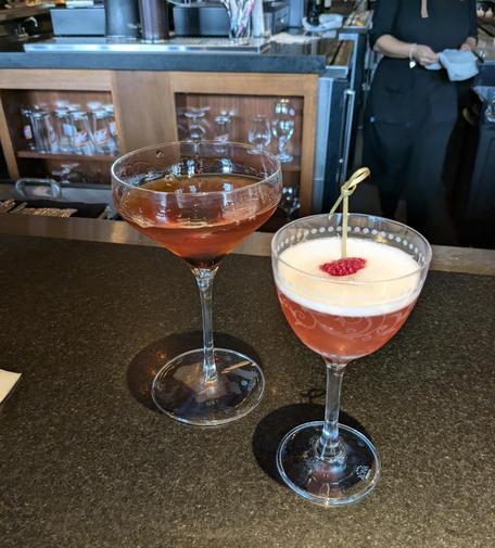 Two cocktails on a bar: a manhattan in a sleek coupe and a frothy pink gin cocktail in a etched cocktail glass.